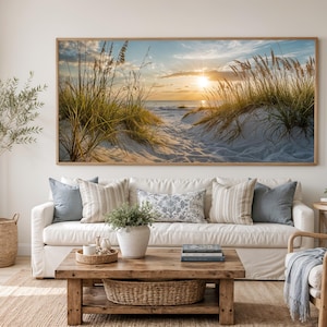 May include: A large rectangular wall art print featuring a coastal sunset scene with golden light, sand dunes, and tall beach grass. The artwork is displayed in a thin wooden frame above a white sofa with decorative pillows in a living room setting.