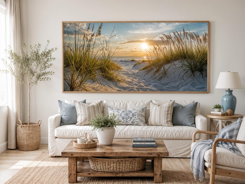 May include: A large rectangular wall art print featuring a coastal sunset scene with golden light, sand dunes, and tall beach grass. The artwork is displayed in a thin wooden frame above a white sofa with decorative pillows in a living room setting.