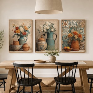 May include: A set of three framed wall art prints featuring still life compositions of terracotta pottery, blue pitchers, and floral arrangements. The artwork displays vibrant orange and white flowers alongside patterned tiles in a neutral color palette.