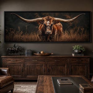 Op de afbeelding: Een panoramische wandkunstprint met een longhorn-koe met grote, wijd uitlopende hoorns in een veld met droog, goudbruin gras. Het dier met een donkere, getextureerde vacht is ingelijst in een zwarte lijst aan een donkerbruine muur.