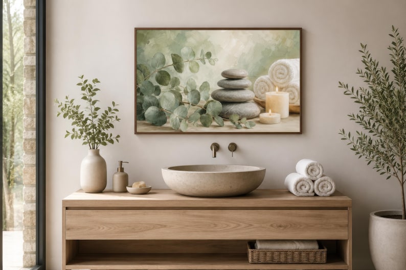 May include: A rectangular wall art print featuring a stack of smooth grey stones, green eucalyptus branches, white rolled towels, and two lit cream-coloured candles. The artwork is displayed in a dark brown frame above a wooden bathroom vanity.