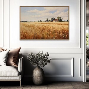 a living room with a large painting of a field of golden wheat hanging on the wall. The painting is framed in a wooden frame and is the focal point of the room. The living room is furnished with a couch, a vase of flowers, and a dining table with chairs. The overall atmosphere of the room is calm and inviting, with the painting adding a touch of nature and warmth to the space.