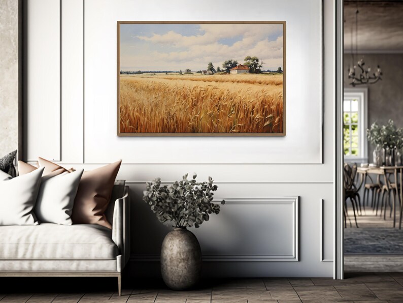a living room with a large painting of a field of golden wheat hanging on the wall. The painting is framed in a wooden frame and is the focal point of the room. The living room is furnished with a couch, a vase of flowers, and a dining table with chairs. The overall atmosphere of the room is calm and inviting, with the painting adding a touch of nature and warmth to the space.