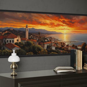 a large framed painting of a picturesque coastal town at sunset, with a lighthouse and mountains in the background. The painting is hung on a wall above a wooden dresser, which has a lamp, a stack of books, and a clock on it.