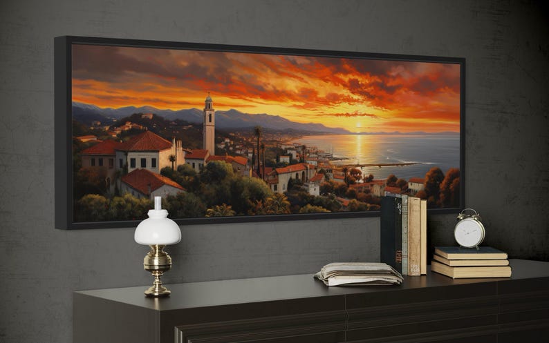 a large framed painting of a picturesque coastal town at sunset, with a lighthouse and mountains in the background. The painting is hung on a wall above a wooden dresser, which has a lamp, a stack of books, and a clock on it.