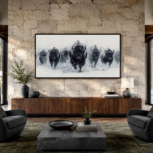 May include: A rectangular wall art print featuring a black and white illustration of a herd of bison running forward. The artwork is displayed in a thin black frame and mounted on a light-colored stone wall above a long wooden media console.