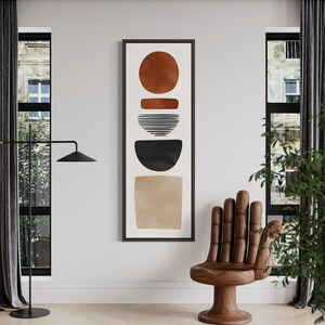 May include: Abstract art print featuring a vertical composition of geometric shapes in shades of brown, black, and beige. The artwork is framed in a dark brown frame and hangs on a white wall. A wooden hand-shaped chair is in the foreground.