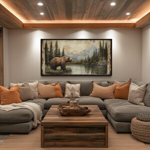 a cozy living room with a large, comfortable gray sectional sofa adorned with orange and beige throw pillows. Above the sofa, a large painting of a bear in a forest setting hangs on the wall. The room features a wooden ceiling with recessed lighting, and a wooden coffee table in the center of the space.