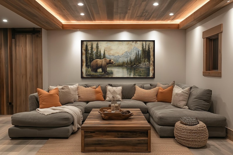 a cozy living room with a large, comfortable gray sectional sofa adorned with orange and beige throw pillows. Above the sofa, a large painting of a bear in a forest setting hangs on the wall. The room features a wooden ceiling with recessed lighting, and a wooden coffee table in the center of the space.