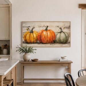 May include: A rectangular wall art piece featuring a painting of three pumpkins in yellow, orange, and muted green, set against a light-colored wooden plank background, enclosed in a thin, light-toned wooden frame hanging on a plain wall.
