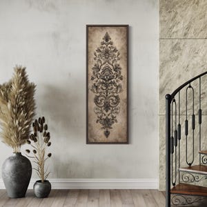 May include: A framed, vertical artwork featuring a detailed, symmetrical damask design in shades of gray and black. The artwork is set against a beige background and framed in a dark brown border. The artwork is displayed on a light-colored wooden floor.