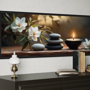A framed painting depicting a serene arrangement of white flowers, gray stones, and a lit candle on a wooden surface, with a lamp and books nearby.
