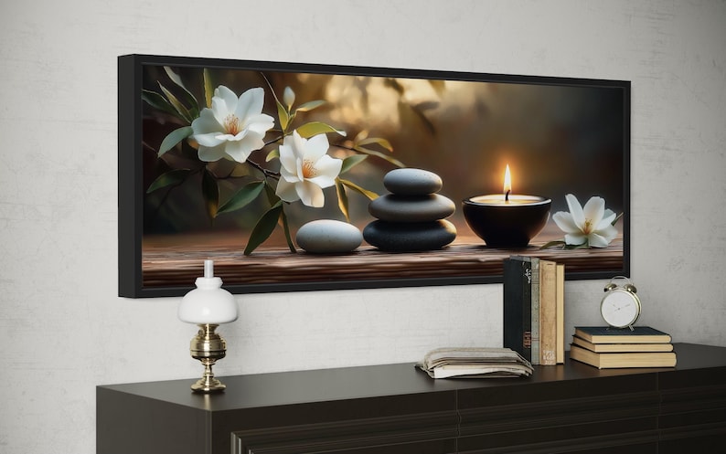 A framed painting depicting a serene arrangement of white flowers, gray stones, and a lit candle on a wooden surface, with a lamp and books nearby.