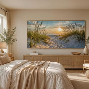 May include: A large rectangular wall art print featuring a sunset over a sandy beach with dune grass, displayed above a long wooden sideboard in a bedroom. The room includes a bed with beige bedding, a lamp, a chair, and a woven ottoman.