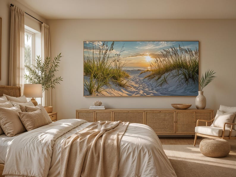 May include: A large rectangular wall art print featuring a sunset over a sandy beach with dune grass, displayed above a long wooden sideboard in a bedroom. The room includes a bed with beige bedding, a lamp, a chair, and a woven ottoman.