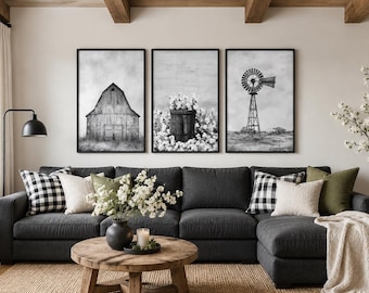May include: A set of three black and white framed wall art prints featuring a rustic barn, a potted flower arrangement, and a metal windmill. The prints are displayed in thin black frames and hung in a horizontal row above a dark gray sectional sofa.