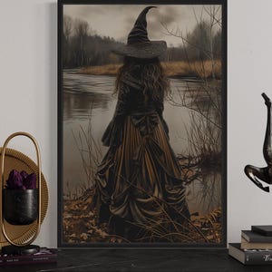 a framed artwork depicting a woman in a witch's hat standing by a lake, with a statue of a horse next to it. The artwork is displayed on a shelf, accompanied by a candle holder, a book, and a small sculpture.