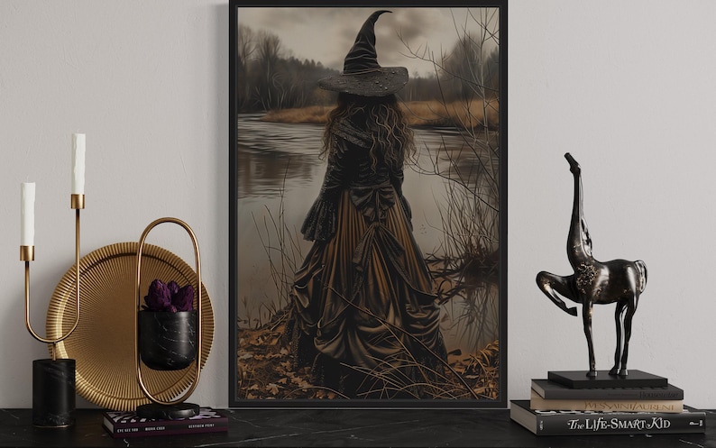 a framed artwork depicting a woman in a witch's hat standing by a lake, with a statue of a horse next to it. The artwork is displayed on a shelf, accompanied by a candle holder, a book, and a small sculpture.