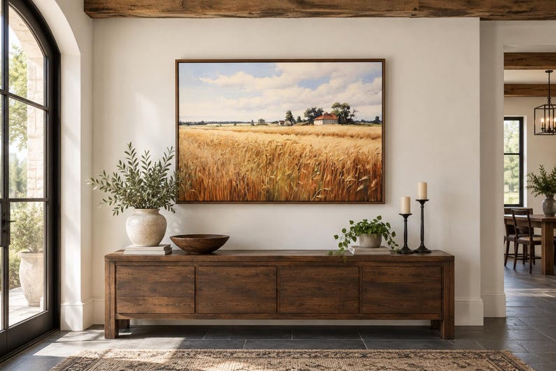 May include: A rectangular landscape painting featuring a golden wheat field under a blue sky with white clouds, displayed in a thin brown frame above a long, dark wooden console table with four cabinet doors and a smooth, flat top surface.