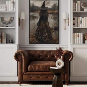a cozy living room with a brown leather armchair, a wooden side table, and a bookshelf filled with books and decorative items. A large painting of a woman in a long black dress is hanging on the wall above the armchair, and a vase of flowers is placed on the side table.