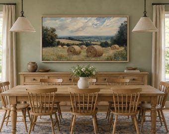 Op de afbeelding: Een rechthoekig landschapsschilderij van een goudkleurig hooiveld met ronde hooibalen onder een bewolkte lucht, ingelijst in licht hout. Het kunstwerk hangt boven een lang houten dressoir en een eettafel met zes houten stoelen.