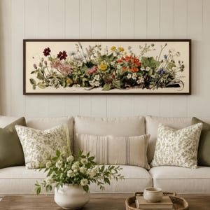Può includere: Una stampa botanica rettangolare incorniciata con una varietà di fiori colorati. L'opera d'arte è appesa sopra un divano color crema adornato con cuscini verdi e fantasia, creando un'atmosfera accogliente.