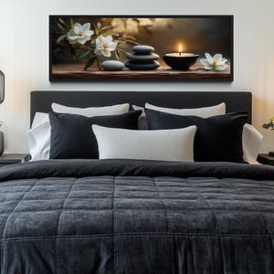 a serene and cozy bedroom scene, featuring a large bed with a gray comforter and a black headboard, flanked by two pendant lights. Above the bed, there is a framed artwork depicting a still life arrangement of flowers and stones. The room also includes a nightstand on each side of the bed, with a vase of flowers and a candle providing a warm and inviting atmosphere.