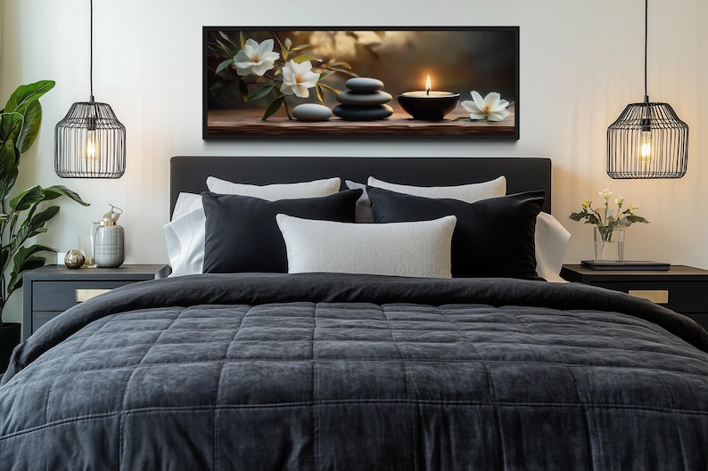 a serene and cozy bedroom scene, featuring a large bed with a gray comforter and a black headboard, flanked by two pendant lights. Above the bed, there is a framed artwork depicting a still life arrangement of flowers and stones. The room also includes a nightstand on each side of the bed, with a vase of flowers and a candle providing a warm and inviting atmosphere.