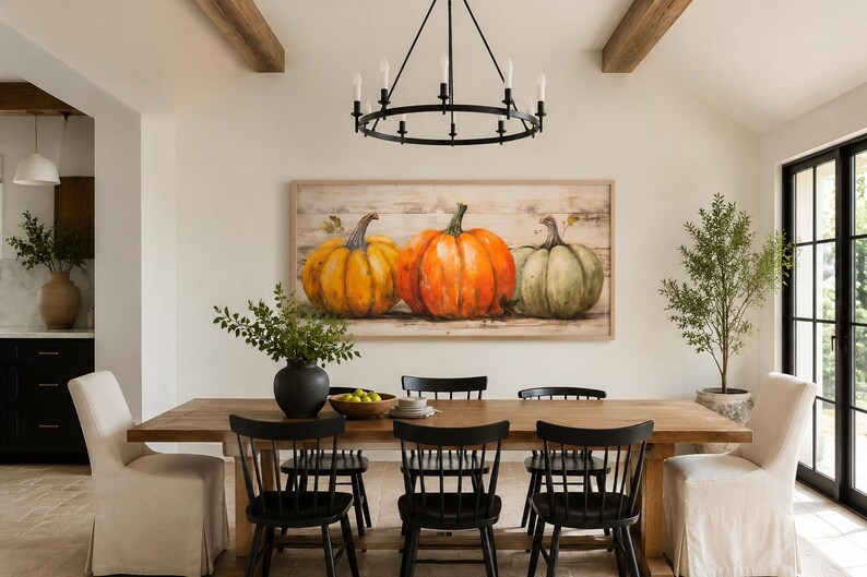 May include: A rectangular wall art piece featuring a painting of three pumpkins in yellow, orange, and green hues, set against a light wood-plank background. The artwork is framed in a light-colored wood frame and displayed above a wooden dining table.