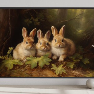 A framed painting of three rabbits sitting on a bed of leaves and branches, surrounded by a forest setting.