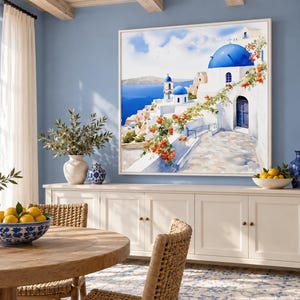 May include: A square framed wall art print depicting a Mediterranean coastal scene with white buildings, blue domes, and orange flowers. The artwork is displayed on a light blue wall above a long white wooden sideboard cabinet.