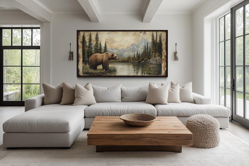 a large painting of a bear in a forest setting, hanging on the wall above a white sectional sofa with beige pillows. The sofa is positioned in front of a wooden coffee table, and there is a large window in the background that provides a view of the outdoors.