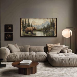 a cozy living room with a large painting of a bear in a forest scene hanging on the wall. The room features a beige sofa, a round wooden coffee table, and a floor lamp. The walls are painted in a neutral gray color, and the room is filled with bookshelves, creating a warm and inviting atmosphere.