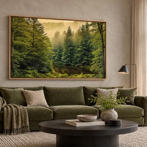 May include: A large rectangular framed wall art print depicting a dense forest with tall green pine trees and a misty, golden-lit background. The artwork is displayed above a long olive green sofa with textured throw pillows and a blanket.