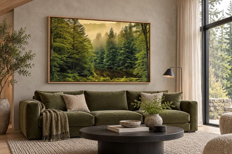May include: A large rectangular framed wall art print depicting a dense forest with tall green pine trees and a misty, golden-lit background. The artwork is displayed above a long olive green sofa with textured throw pillows and a blanket.