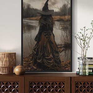 a dark-themed room with a large framed painting of a woman in a long dress standing by a lake, surrounded by trees. The painting is hanging on a wall above a wooden cabinet or dresser, which has a lamp and a vase on top.