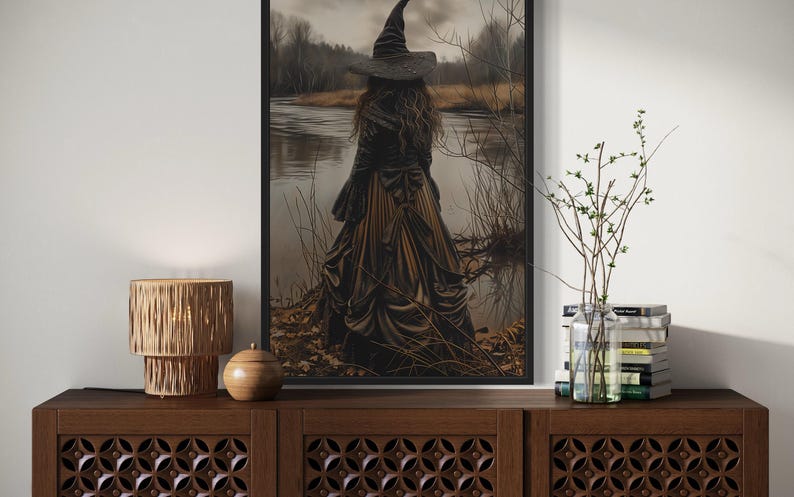 a dark-themed room with a large framed painting of a woman in a long dress standing by a lake, surrounded by trees. The painting is hanging on a wall above a wooden cabinet or dresser, which has a lamp and a vase on top.