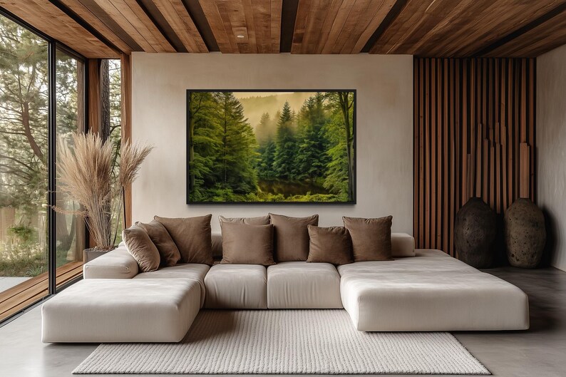 a modern living room with a large window overlooking a forest. The room has a white sofa with brown throw pillows, and a large framed landscape painting hanging on the wall above the sofa.