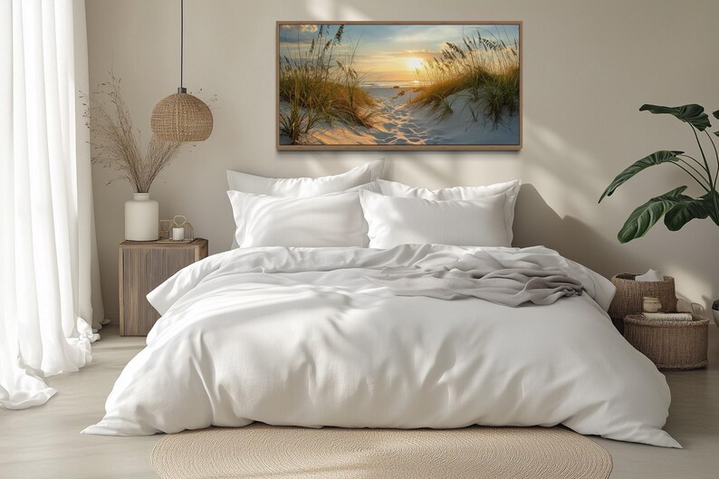 a serene bedroom scene with a large bed, white bedding, and a framed painting of a beach scene above it. The room has a minimalist and modern design, with a large window allowing natural light to fill the space.