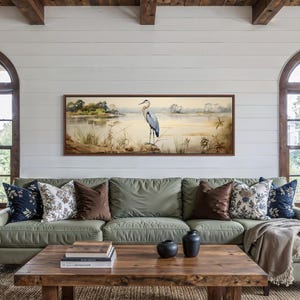 Peut inclure: Salon avec un canapé vert sauge orné de coussins décoratifs. Un tableau de paysage avec un héron est accroché au-dessus. La pièce comprend des fenêtres cintrées et une table basse en bois, créant une ambiance chaleureuse.