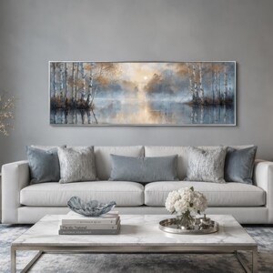 May include: A modern living room with a landscape painting above a white sofa adorned with blue and silver cushions. A marble coffee table displays books and a glass bowl. A vase of flowers sits on a side table, and a lamp provides soft lighting.