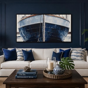 May include: A rectangular wall art print featuring a close-up of a dark blue boat hull, displayed above a white sofa with navy blue and patterned throw pillows. The coffee table holds books titled The Sea and Photographs by the Sea.