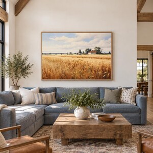May include: A large rectangular landscape painting of a golden wheat field under a blue sky with white clouds, framed in light wood, hanging above a blue sectional sofa, a wooden coffee table, and two brown leather armchairs in a room.