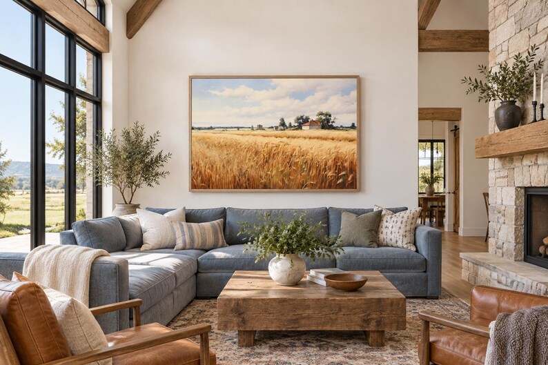 May include: A large rectangular landscape painting of a golden wheat field under a blue sky with white clouds, framed in light wood, hanging above a blue sectional sofa, a wooden coffee table, and two brown leather armchairs in a room.