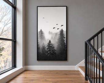 May include: A vertical rectangular wall art piece in a black frame featuring a black and white landscape of a dense pine forest shrouded in thick fog with several birds flying in the sky above the treetops, displayed on a gray interior wall.