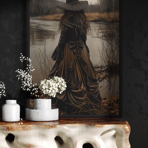 a woman in a long black dress with a pointed hat, standing in front of a painting of a woman in a similar dress, with a body of water and trees in the background.