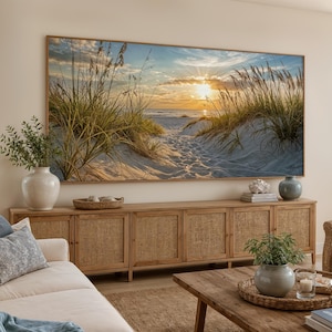 May include: A large rectangular wall art print featuring a sunset over a sandy beach with tall dune grasses in the foreground. The scene depicts a golden sun low on the horizon, casting light over the ocean waves and a path through the dunes.