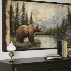 a large painting of a bear standing in a grassy area near a body of water, with mountains in the background. The painting is framed and hangs on a wall above a wooden dresser. On the dresser, there is a lamp, a stack of books, and a clock.