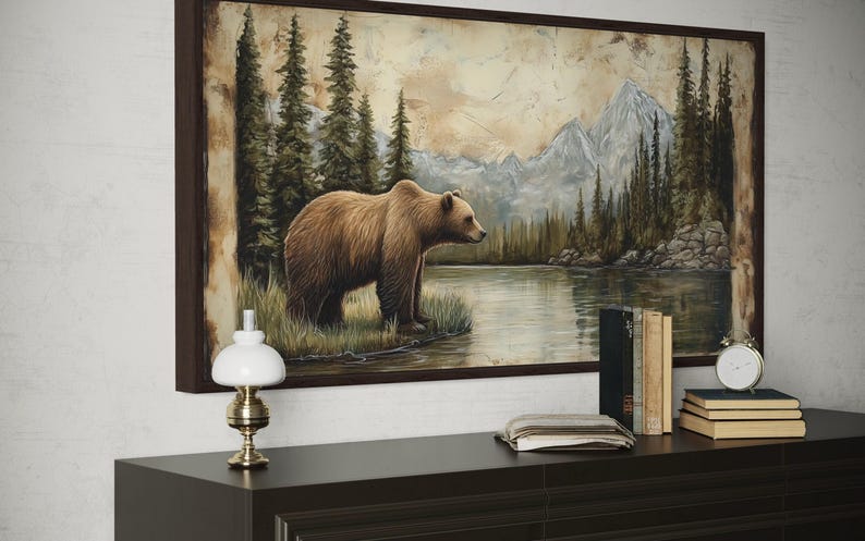 a large painting of a bear standing in a grassy area near a body of water, with mountains in the background. The painting is framed and hangs on a wall above a wooden dresser. On the dresser, there is a lamp, a stack of books, and a clock.