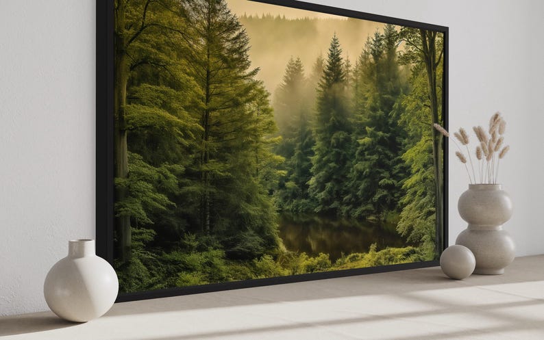 a large framed picture of a forest scene with a lake in the background, placed on a wall. There are two white vases and a potted plant to the right of the picture.